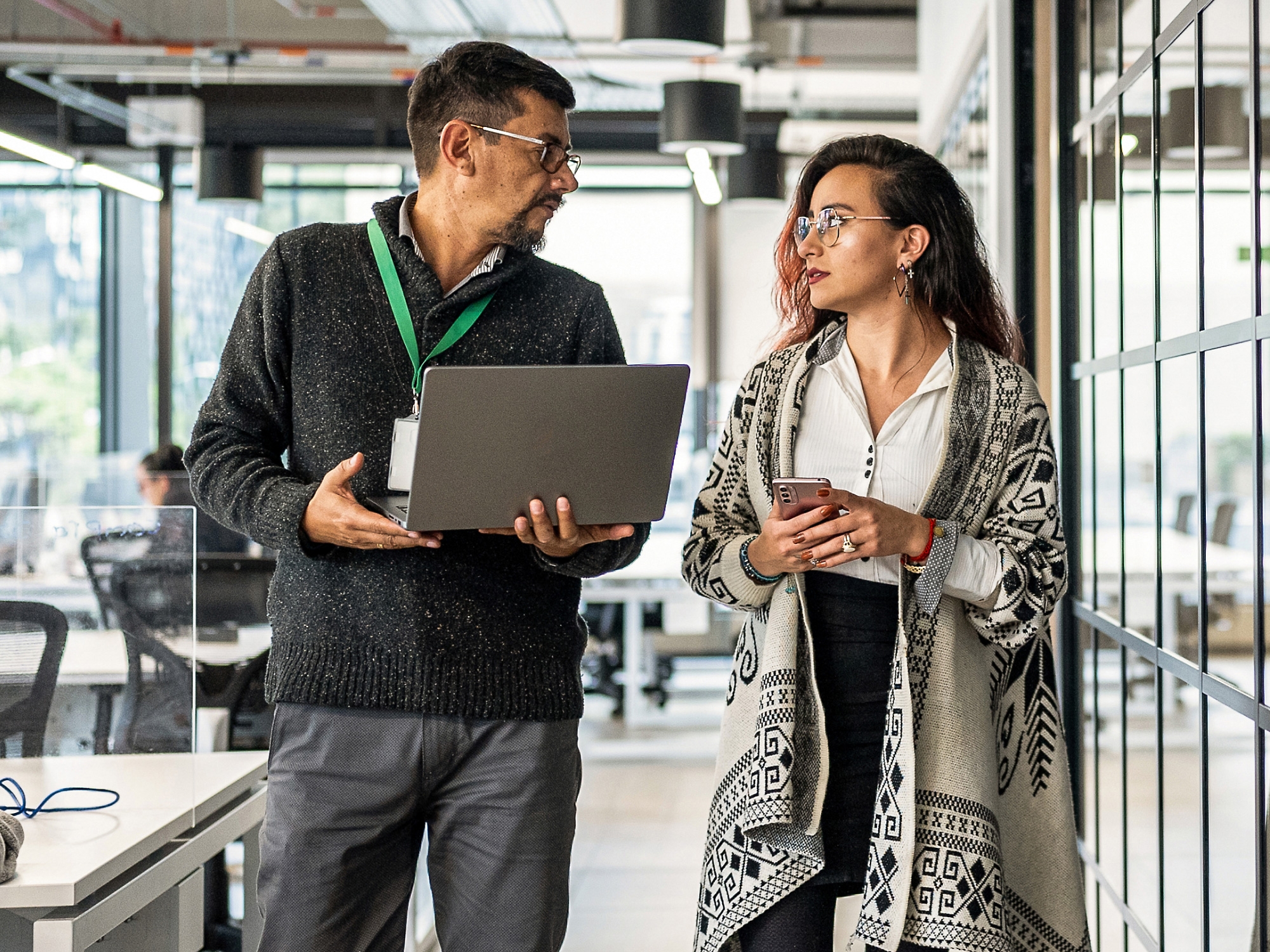 Two professionals discussing, one holding a laptop and the other a smartphone, in a modern office setting.