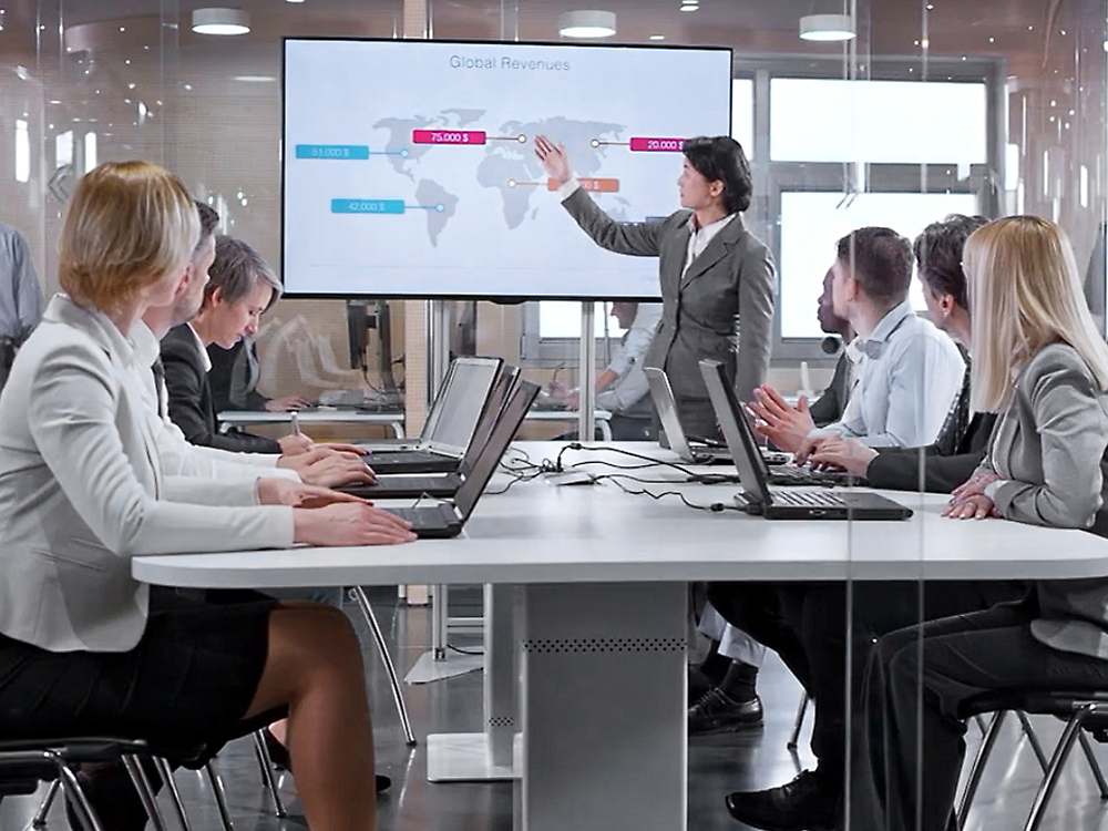 A professional business presentation in a modern office, with a presenter pointing at a screen displaying global revenues