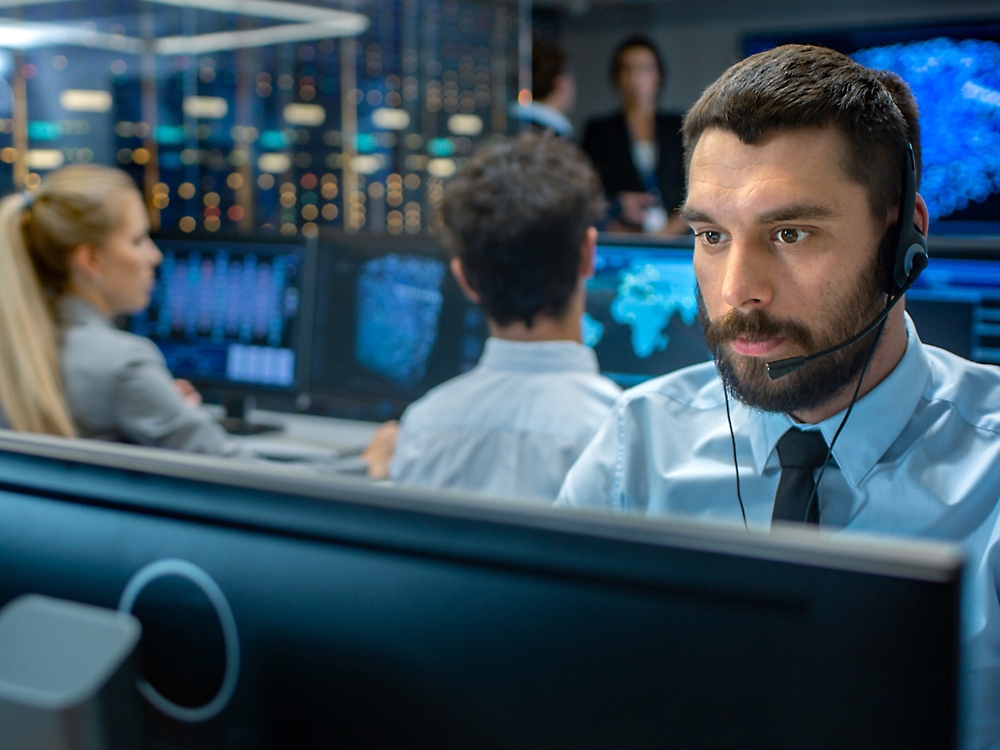 Een man met een headset die op een computer in een drukke controleruimte werkt met collega's en grote schermen waarop gegevens worden weergegeven