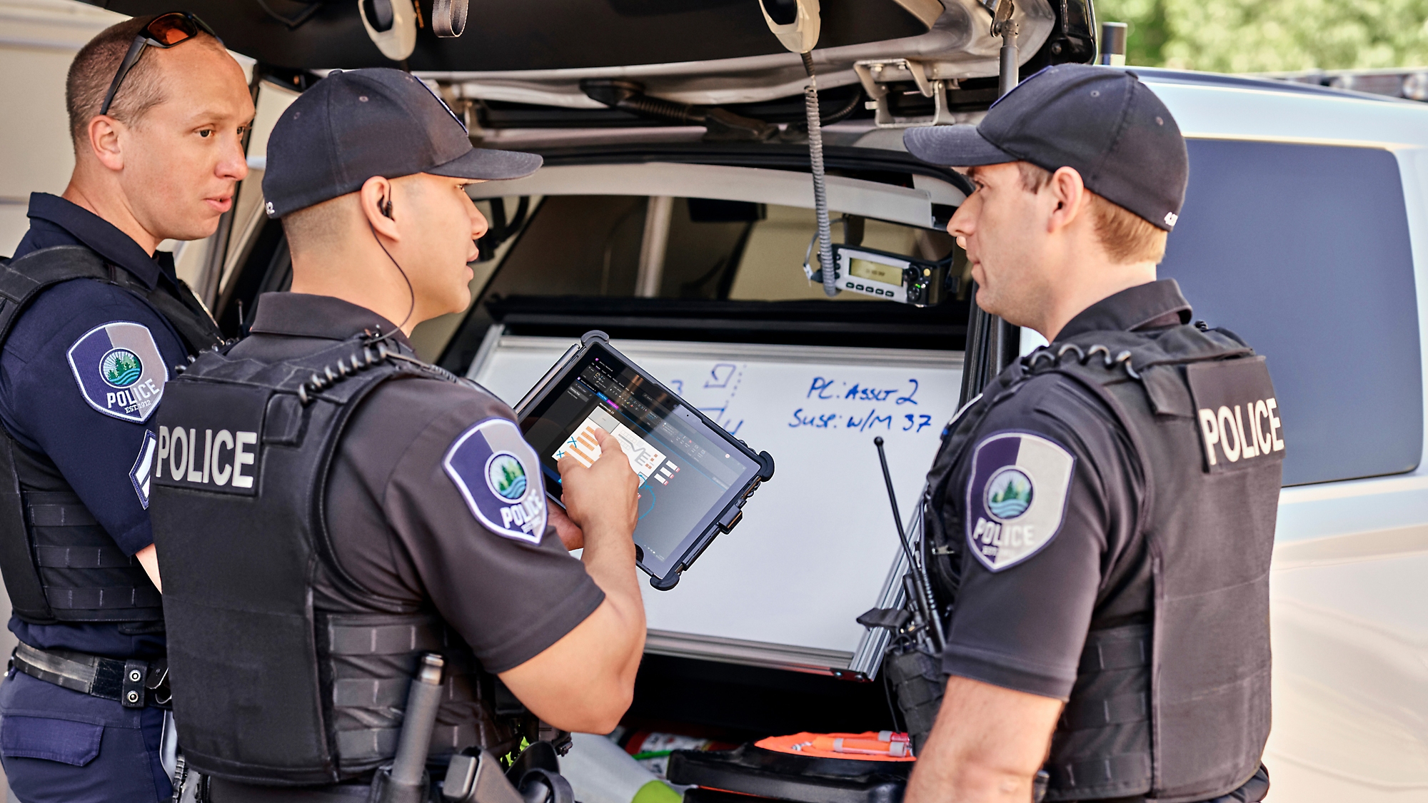 Tres agentes de policía revisan la información de una tableta digital junto a su coche de policía con el maletero abierto