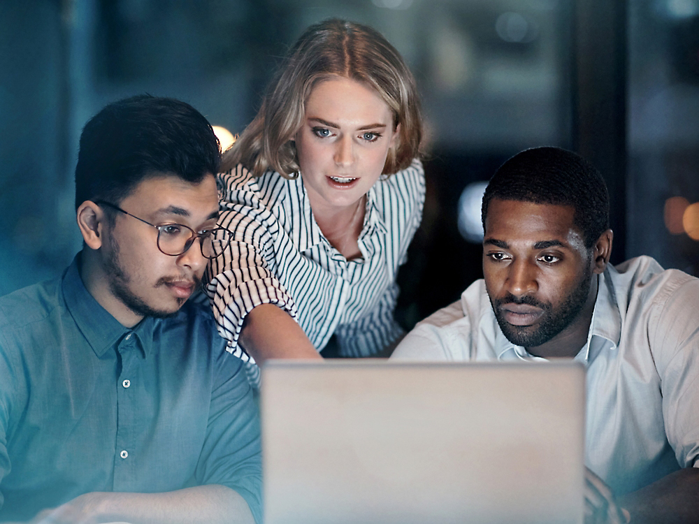 Three colleagues, a woman and two men, focus intently on a laptop screen in a dimly lit office environment.