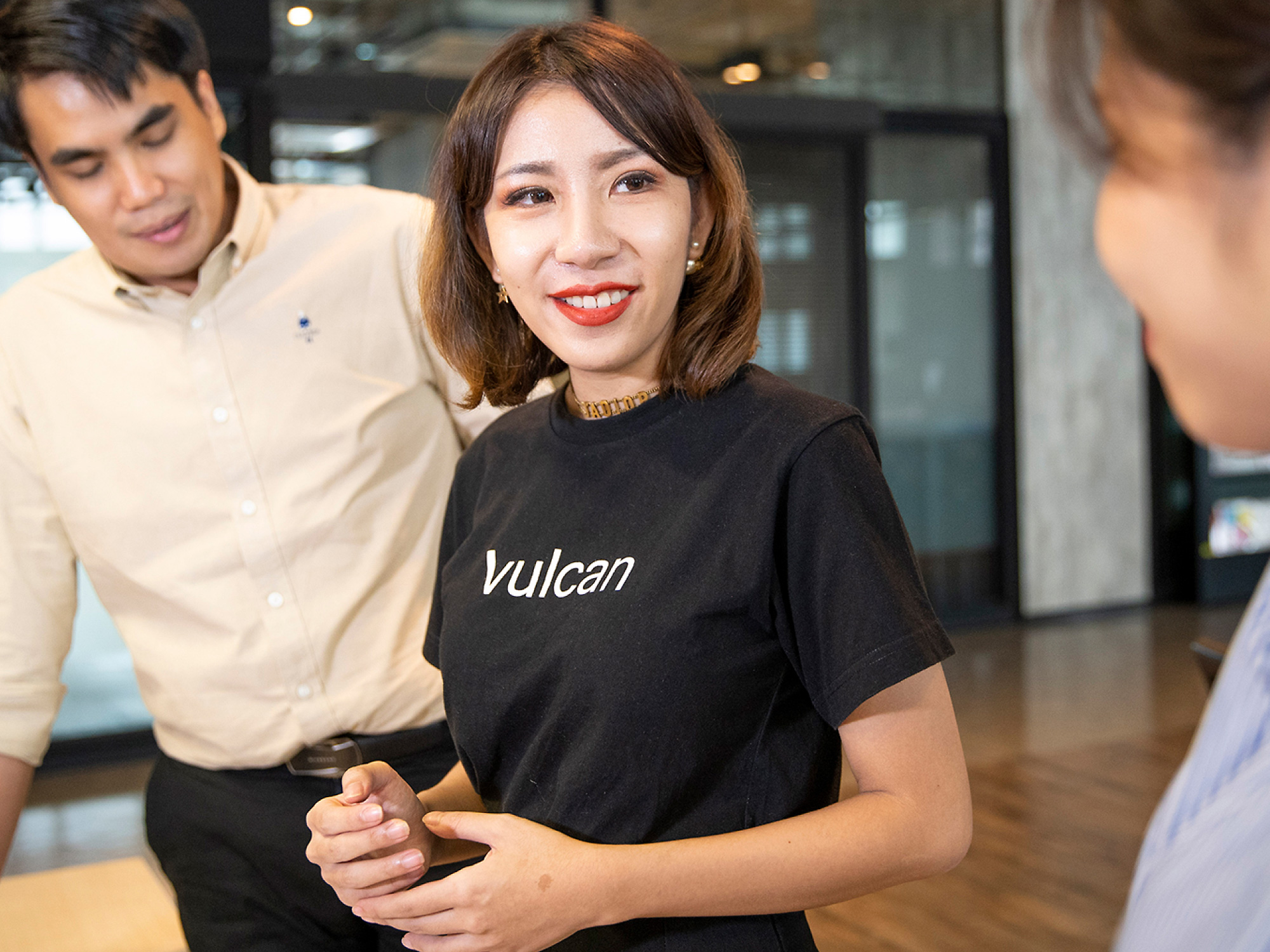 Een jonge vrouw in een zwart t-shirt met het opschrift Vulcan glimlachend naar een collega in een moderne kantooromgeving, met een andere mannelijke collega