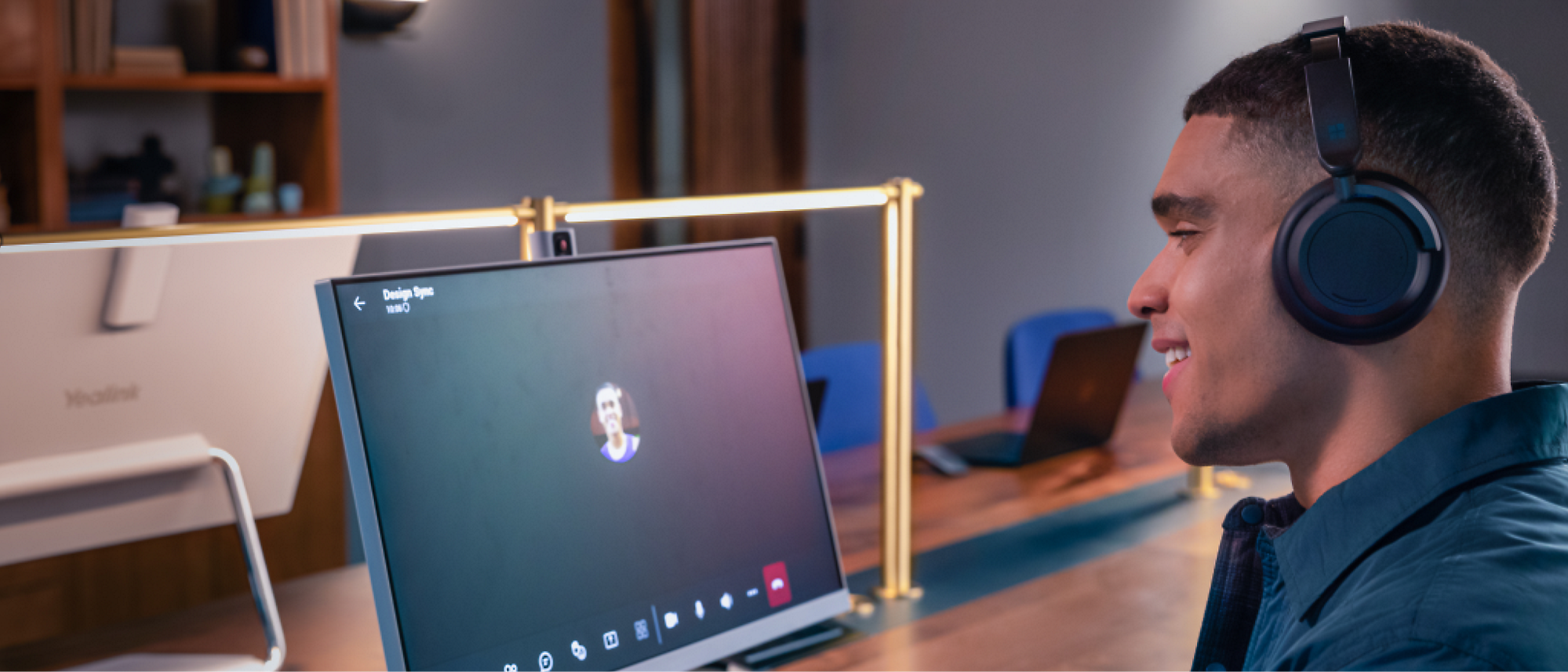 A person wearing headphones is sitting at a desk, engaged in a video call on a computer screen in a modern office setting.