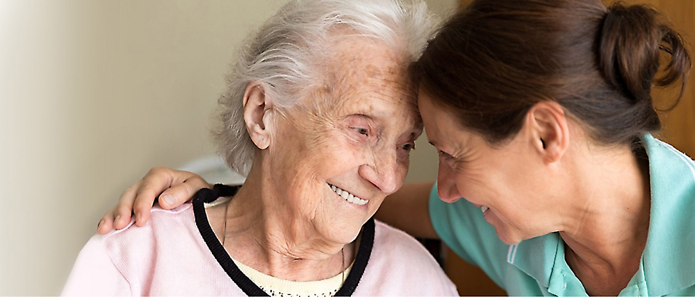Een verzorger die liefdevol lacht naar een oudere vrouw die ook lacht, in een warme binnenomgeving.