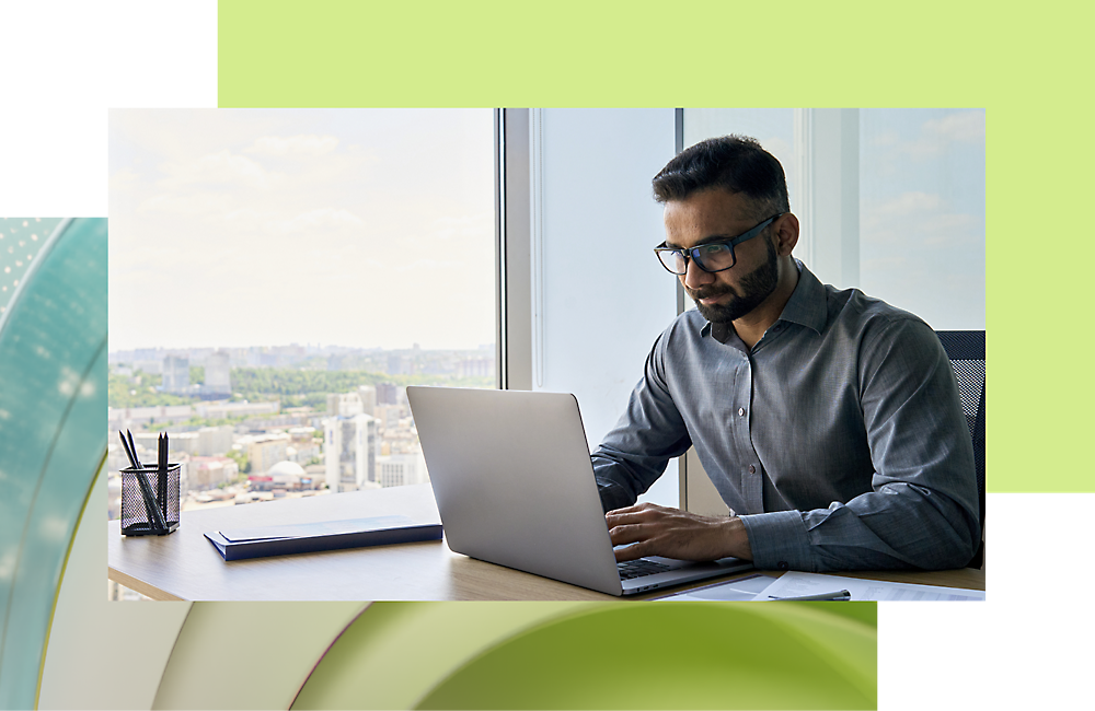 A man wearing glasses, working intently on a laptop at a desk near a window overlooking a cityscape.