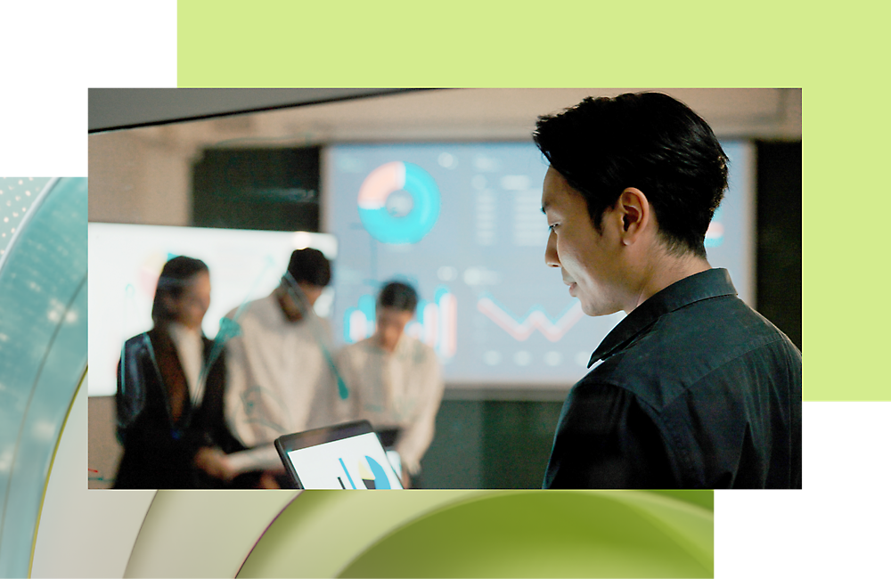 A businessman using a tablet with colleagues discussing data on a digital screen in the background.