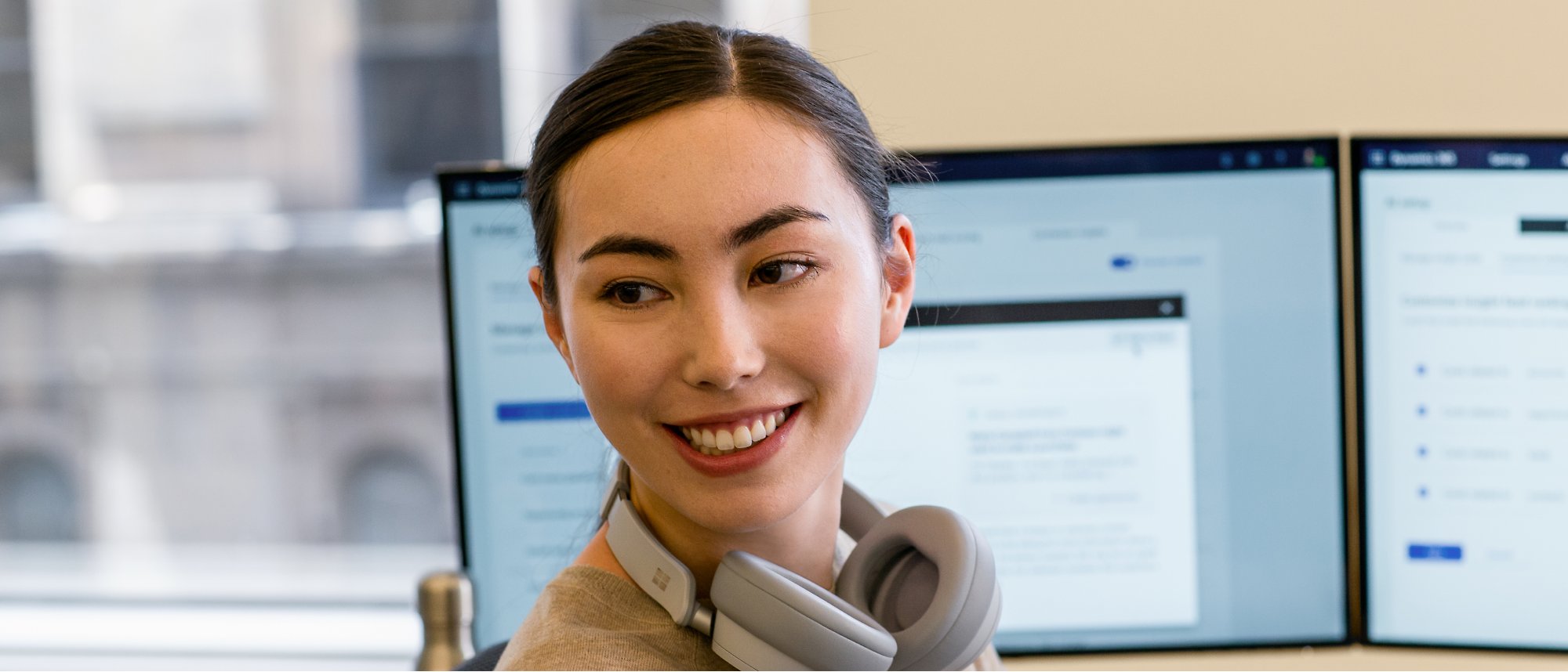 Eine lächelnde Frau, die ihr Headset um den Nacken gelegt hat und an mehreren Monitoren arbeitet.