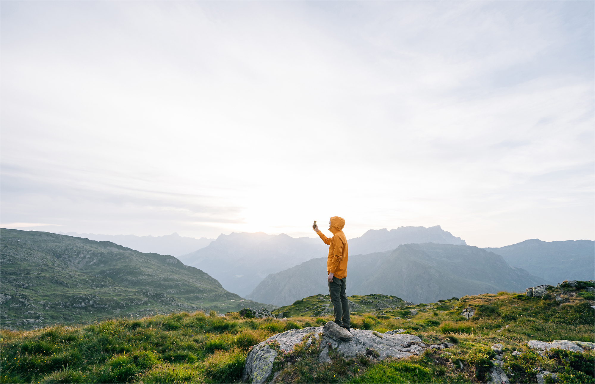 A person stands on a rocky hilltop in a mountainous landscape, holding up a mobile