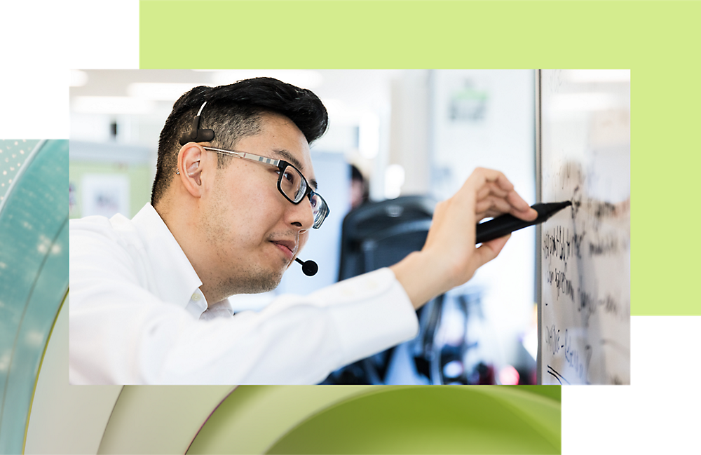 Un homme portant des lunettes et un casque écrit sur un tableau blanc dans un environnement de bureau très actif.