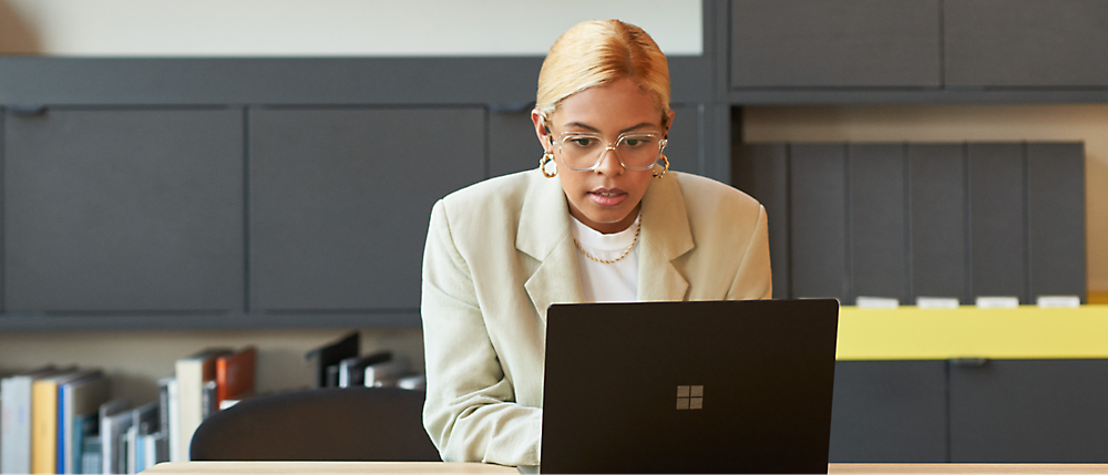 A person with blond hair, wearing glasses and a beige blazer, is working on a laptop in an office setting 