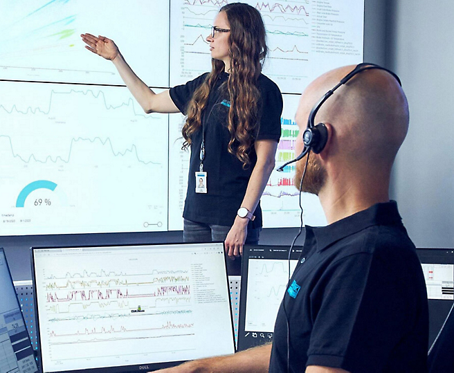 En svejser i beskyttelsesudstyr fokuserer på sit arbejde samtidig med der flyver gnister fra metallet, som han er ved at slibe. En kvinde, der peger på data på en skærm, mens en mand med hovedtelefoner kigger på en computerskærm.