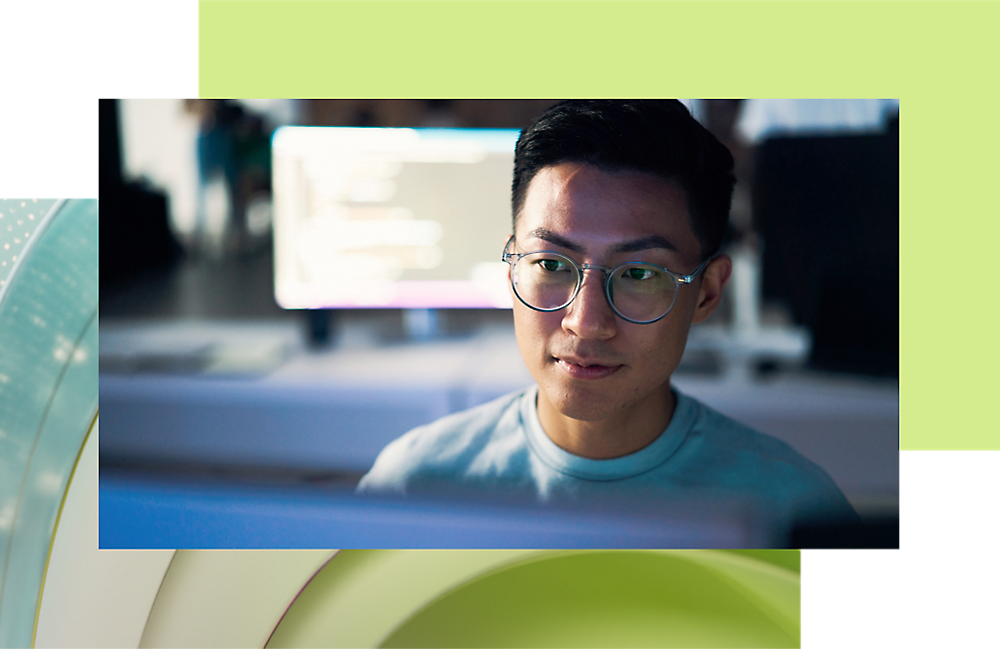 Young asian man wearing glasses, looking at the camera, with a computer screen glowing in the background.