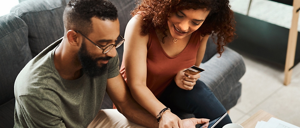 A man and woman sit on a couch looking at a tablet. The woman holds a credit card in her hand.