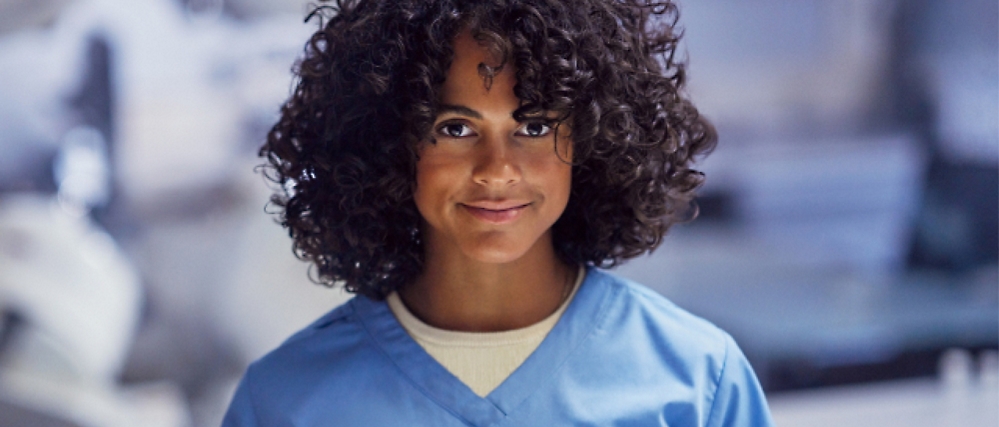 A person with curly hair wearing a light blue scrubs top stands indoors, smiling gently.