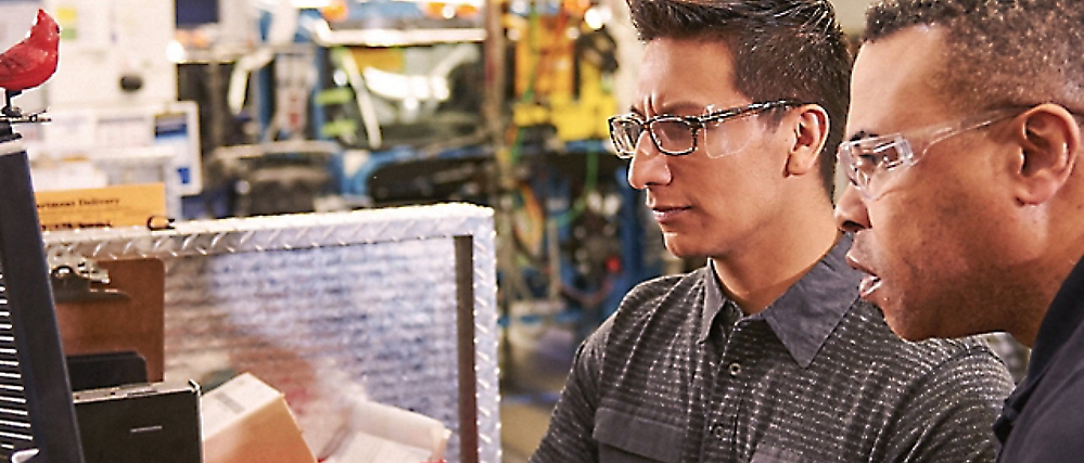 Two men wearing safety glasses focus on a computer screen in an industrial workspace