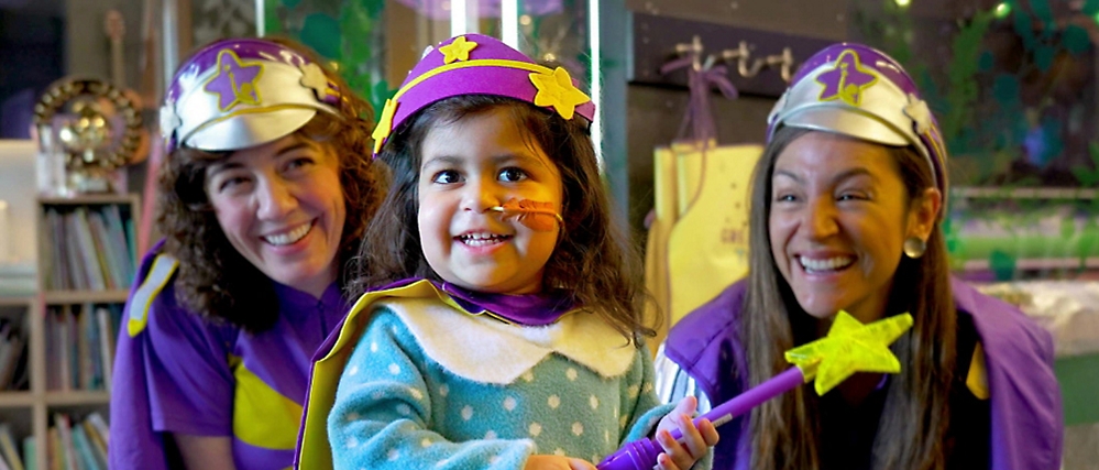 A young child in a blue sweater, wearing a superhero costume with with two other persons