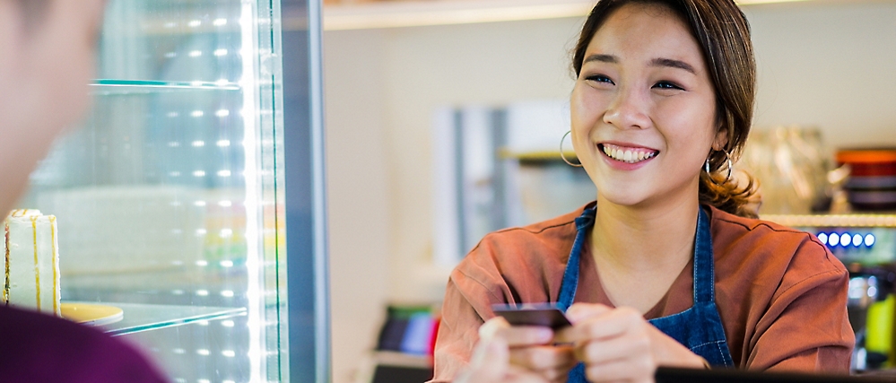 A smiling woman in an apron holds out a credit card for a customer at a counter. 