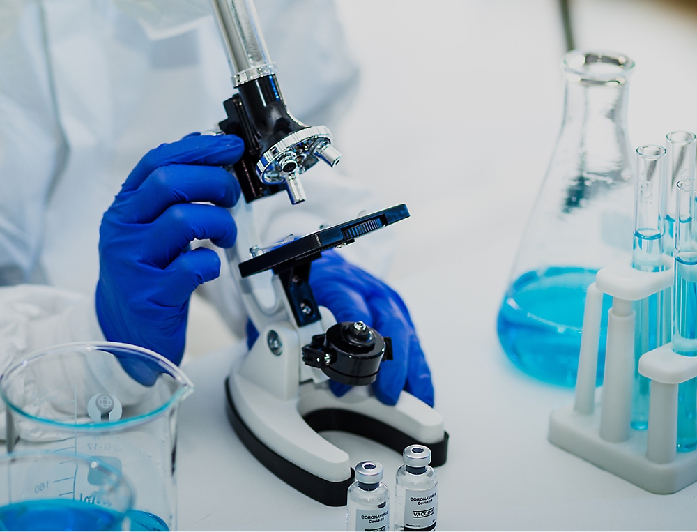 A scientist in blue gloves uses a microscope on a lab table with beakers, test tubes, and small bottles 