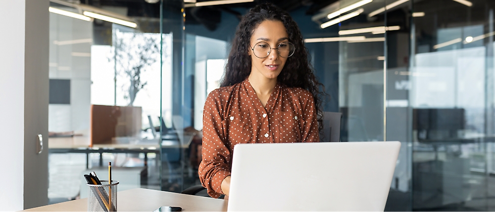 A woman with glasses and curly hair works at a desk with a laptop in a modern office with glass walls.