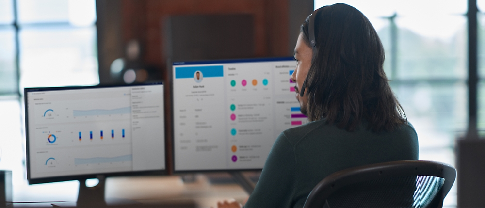 Person sitting at a desk working on two computer monitors displaying graphs and charts in an office setting.