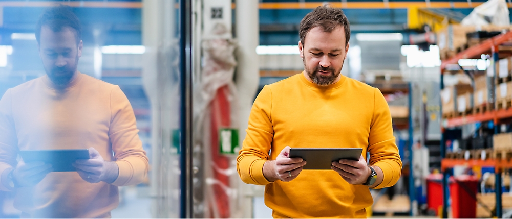 A man in a yellow sweater stands in a warehouse, holding a tablet and looking at the screen.