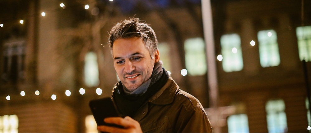 A man standing outdoors at night, smiling while looking at his smartphone. String lights and a building in the background