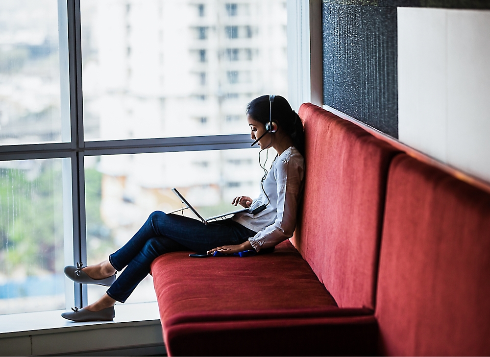 A person wearing a headset sits on a red couch by the window, using a laptop.