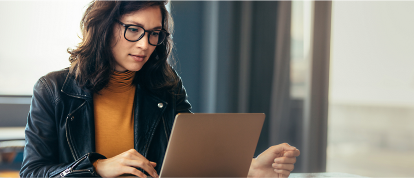 A woman wearing glasses and a black leather jacket sits at a table, working on an open laptop.