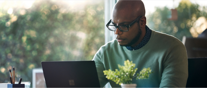 A person wearing glasses and a green sweater works on a laptop at a desk with a small potted plant and a cup of pencils.