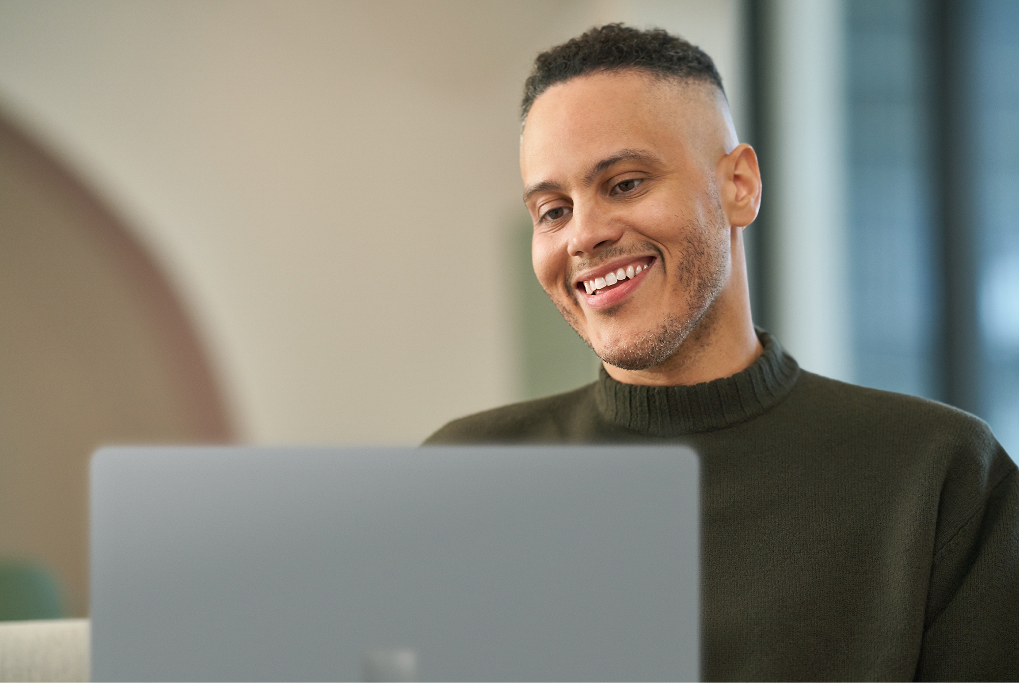 A person with short curly hair, wearing a dark sweater, smiles while using a laptop indoors.