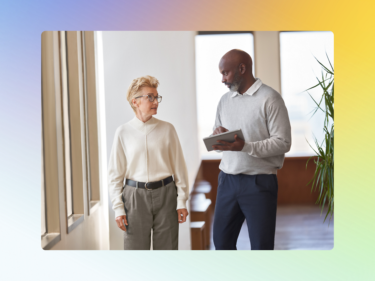 Two people are walking indoors while conversing. One holds a tablet, and the other looks at them.