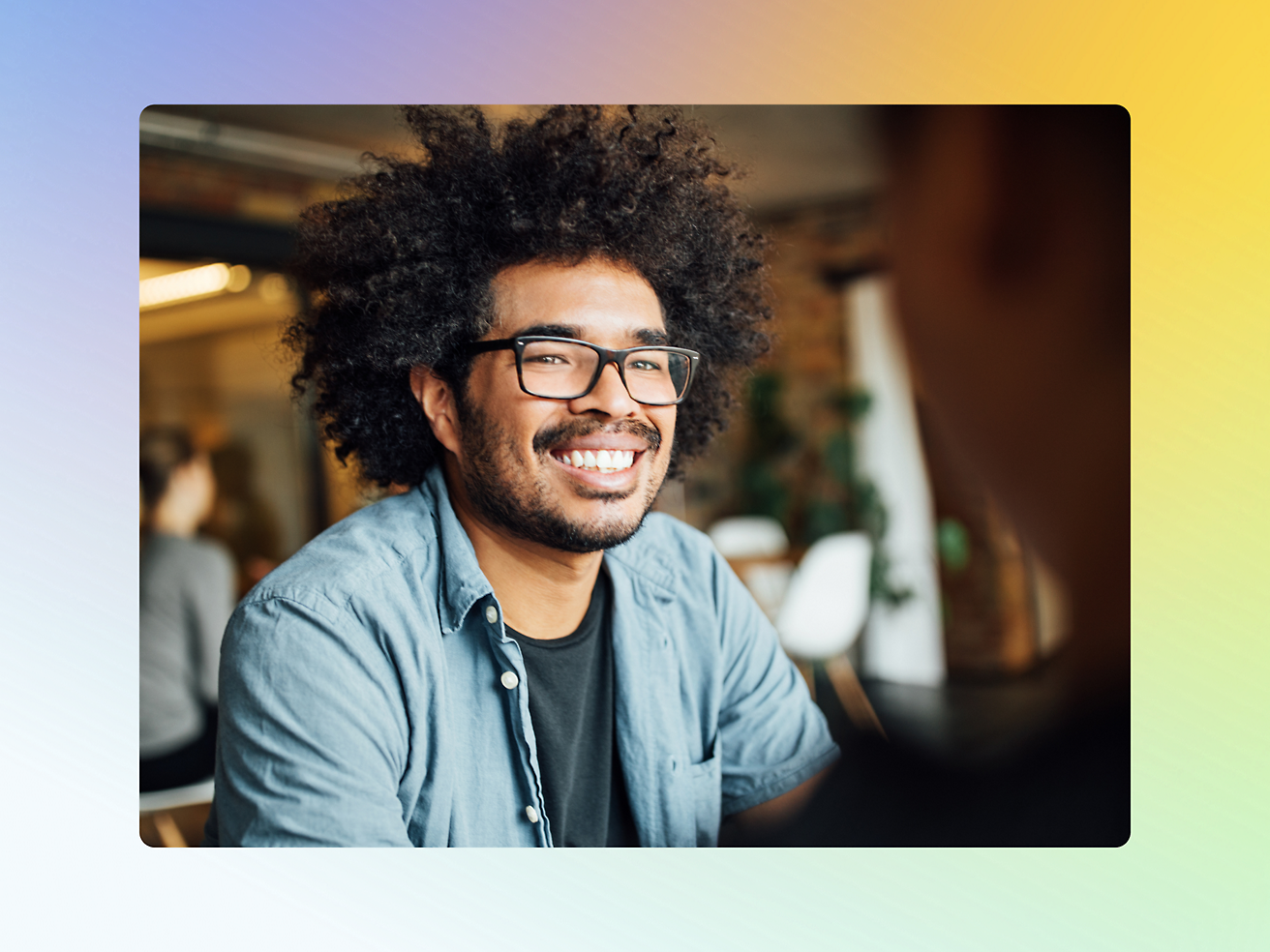 A person with curly hair and glasses smiles while sitting indoors in a casual setting.