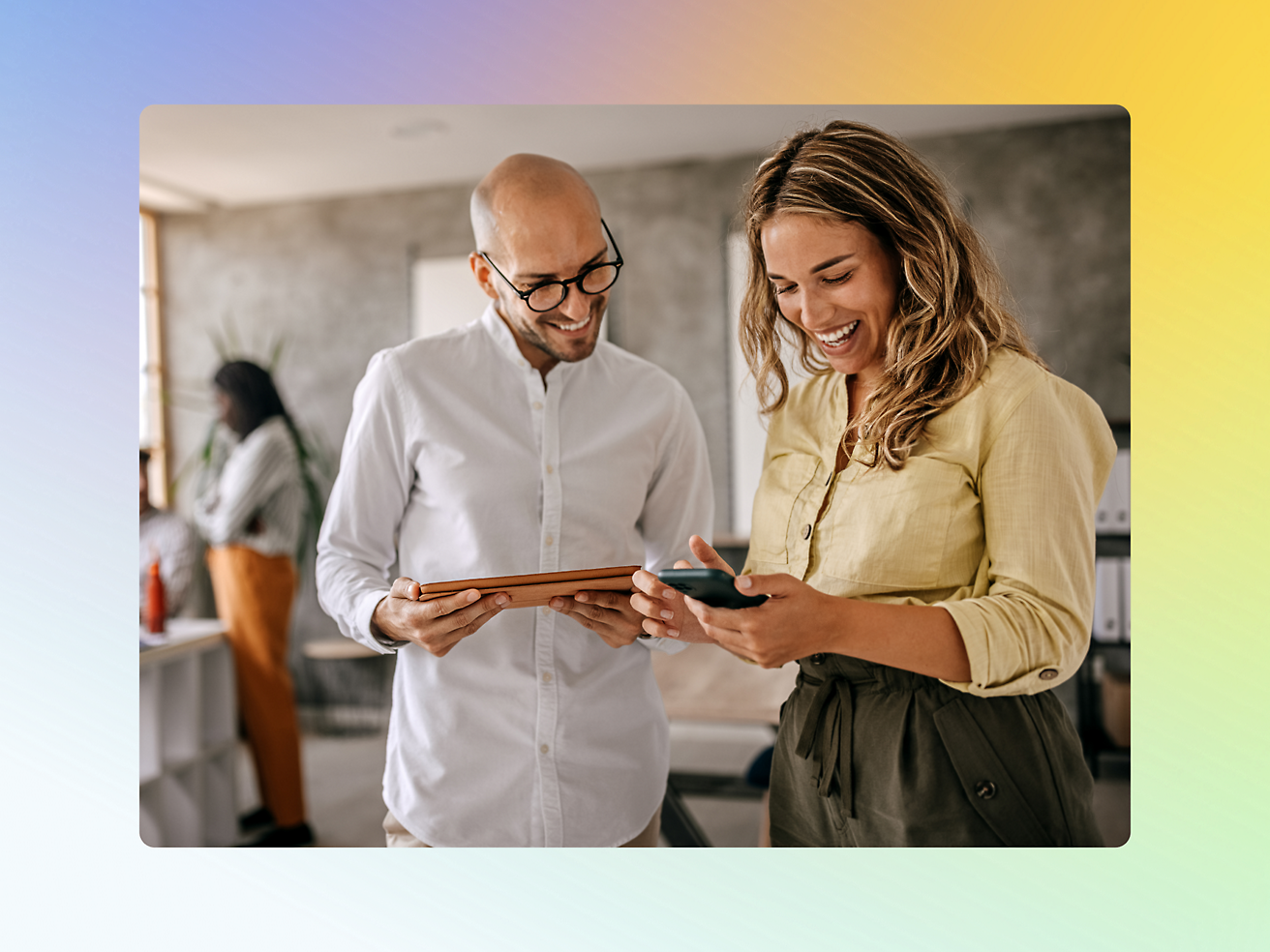 Two colleagues stand together in an office, smiling and looking at a smartphone.