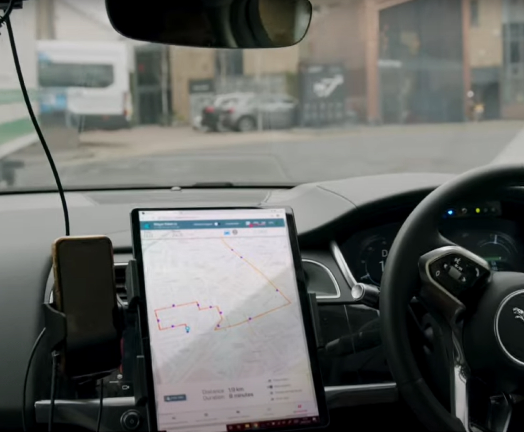 Interior view of a car showing a central touchscreen displaying a map with a route, a mounted smartphone, and the dashboard.