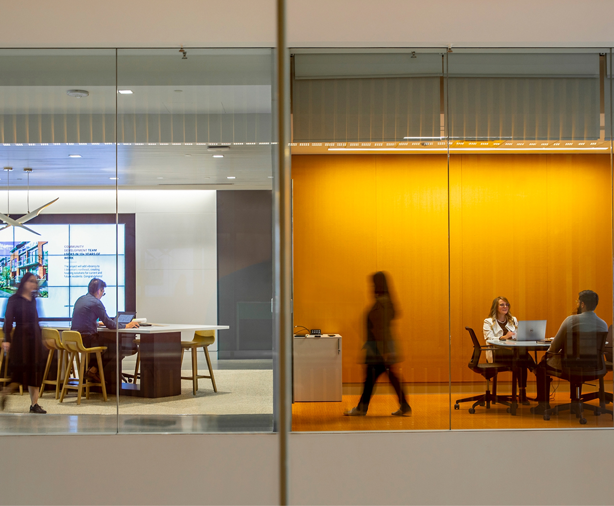 A view through glass walls shows two rooms in an office setting: one with three people at a table with laptops