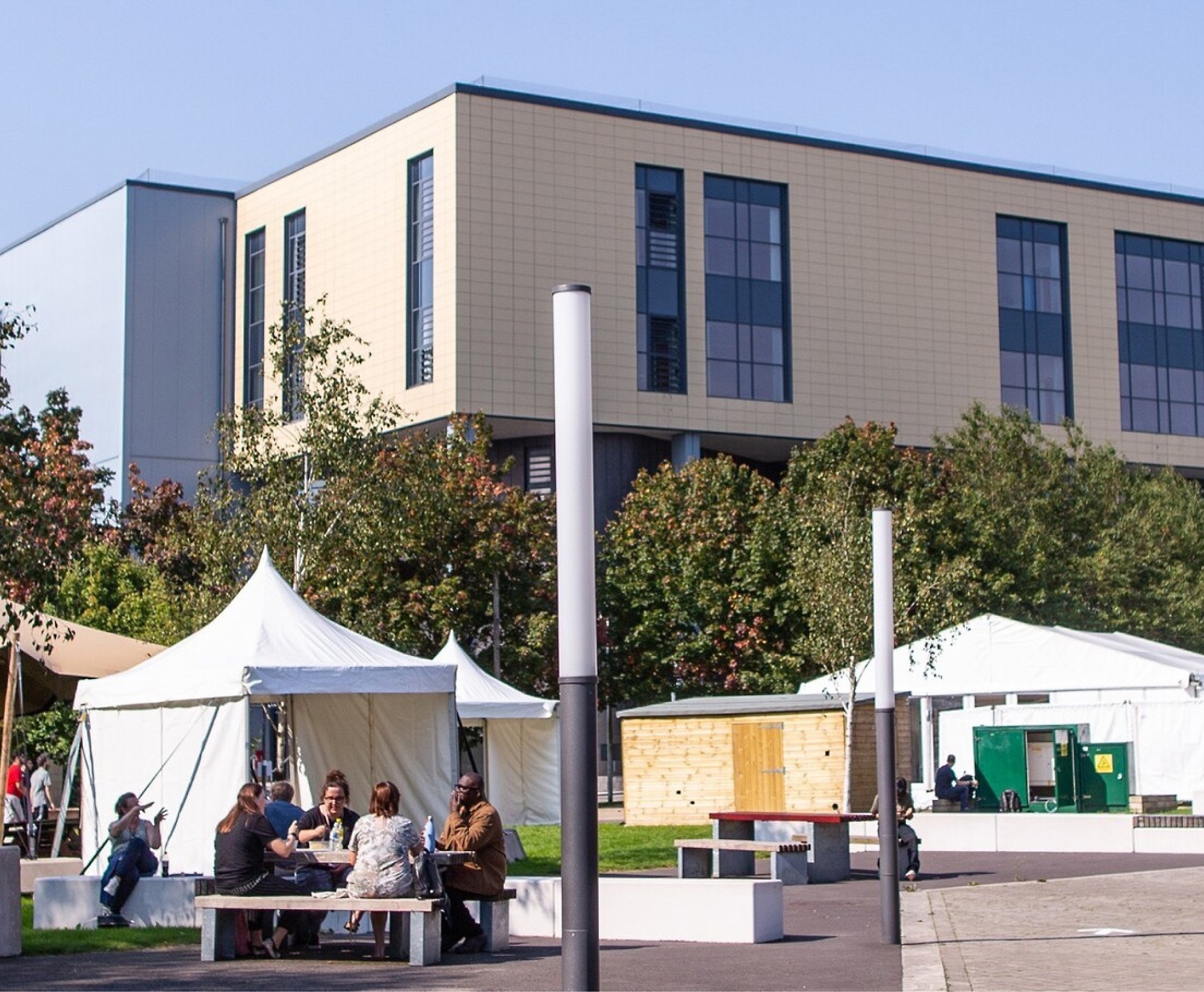 A group of people are sitting and talking at a table in an outdoor space with white tents