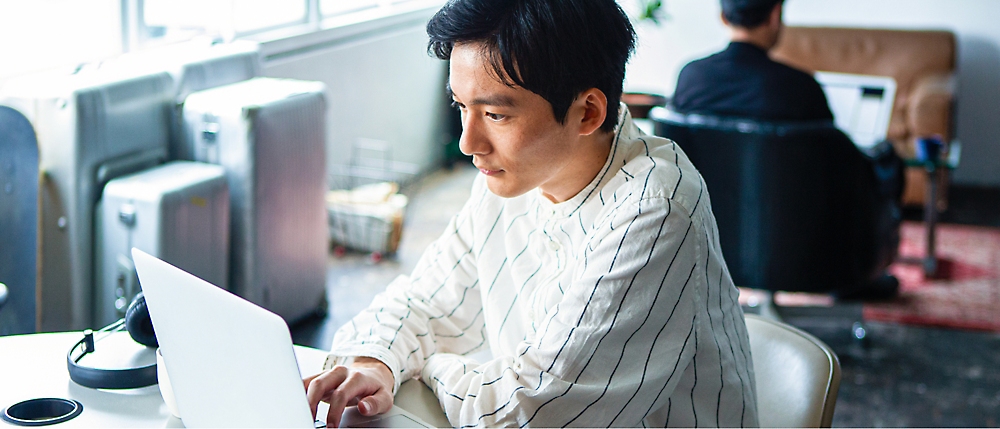 Man in a white shirt working on a laptop at a desk in a well-lit room with another person working in the background.