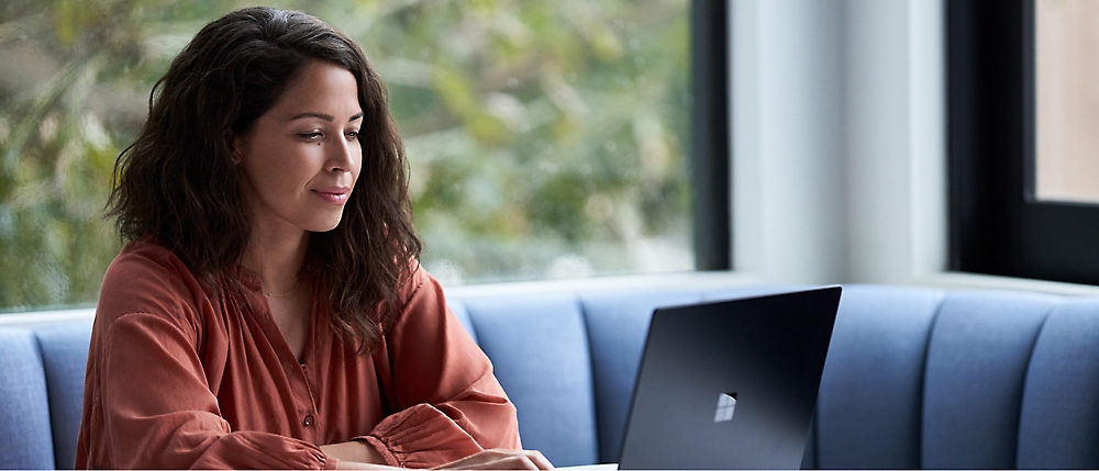 A woman with long brown hair, seated at a blue cushioned bench, works on a laptop near large windows on an overcast day.