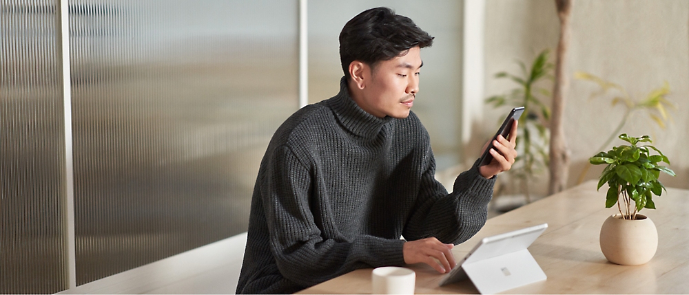 A person in a dark sweater sits at a table using a tablet and holding a smartphone, with a coffee cup and small plant nearby.