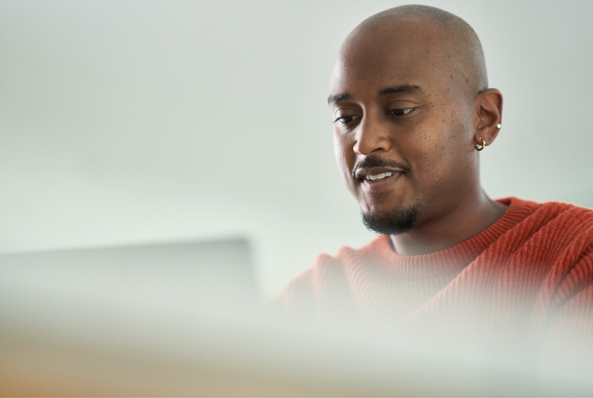 A person with a shaved head and a short beard is looking at a laptop screen, wearing a red sweater.