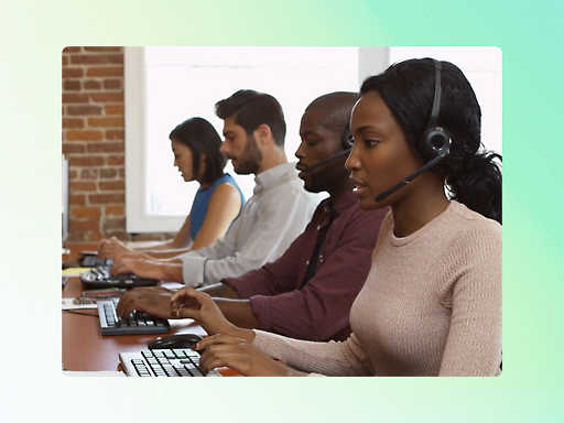 Four individuals wearing headsets are seated in a row, working on computers in an office with brick walls and large windows.