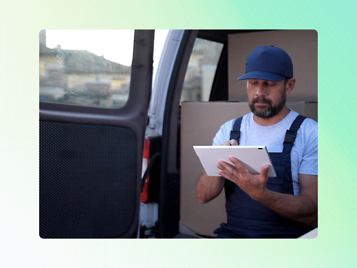 A delivery worker in overalls and a cap is seated in the back of a van, using a tablet computer. Boxes are visible in the background.