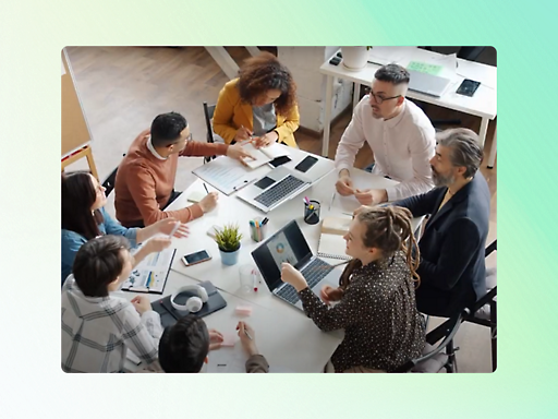 A group of eight people are sitting at a table, collaborating in an office setting. Laptops, notebooks, are on the table.