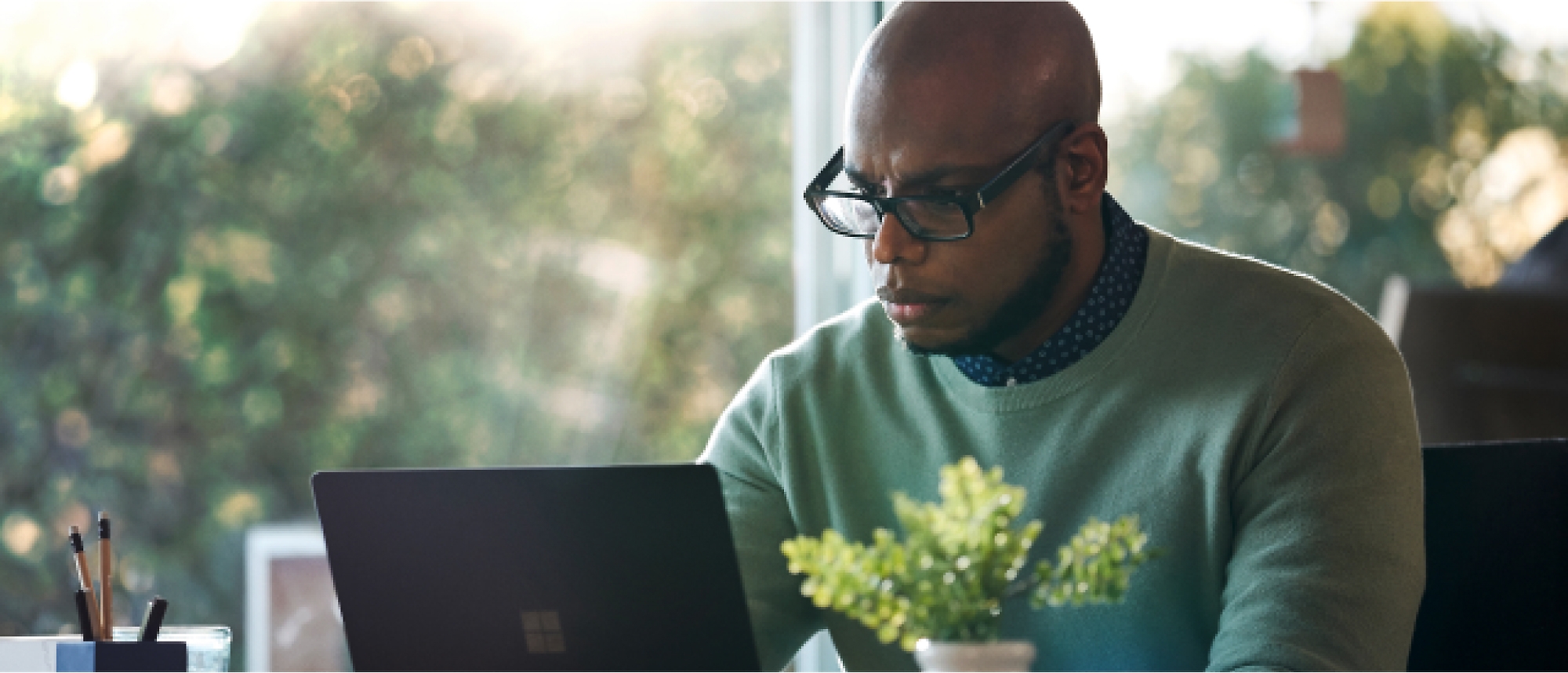 A person with glasses works on a laptop at a desk with a potted plant in the foreground.