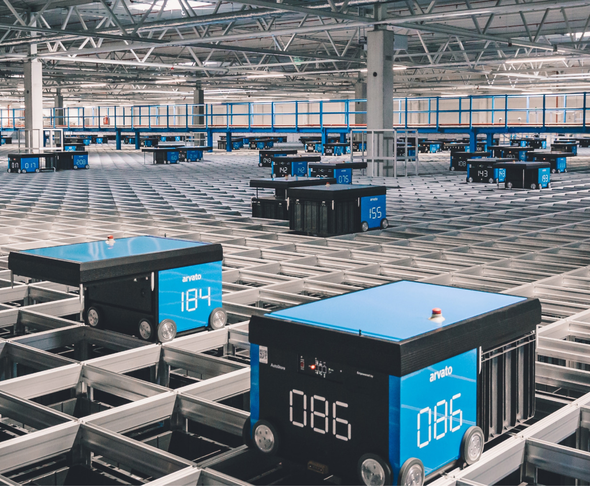 A warehouse filled with numerous blue robotic units on tracks, each displaying numbers. The space is large with a high ceiling and bright lighting.