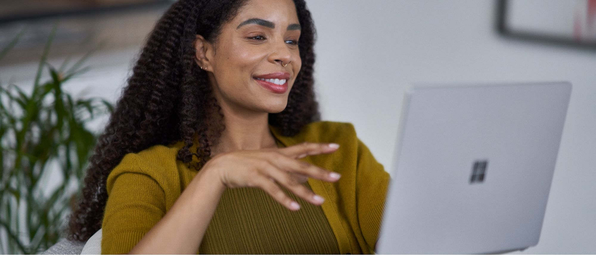 A woman with curly hair, wearing a yellow sweater, gestures while looking at a laptop.