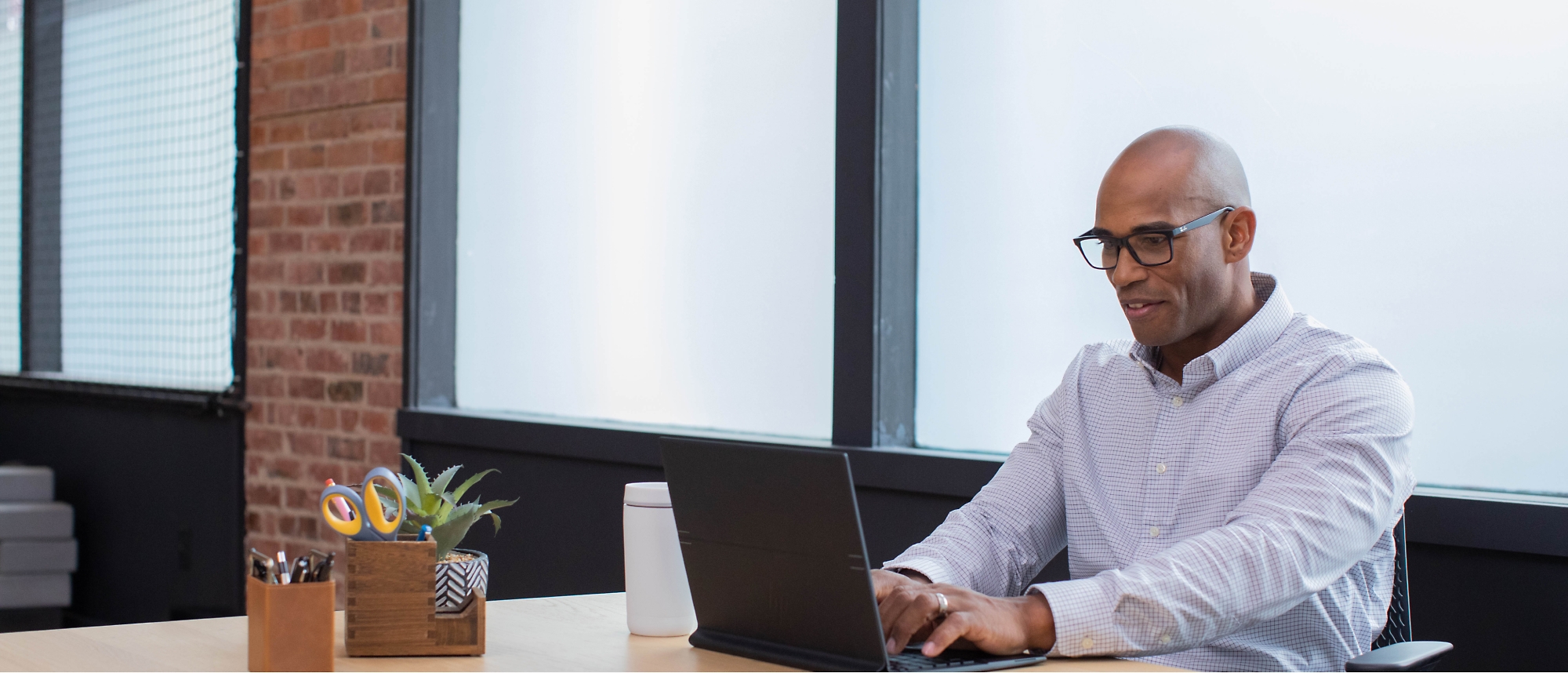 Man in a light blue shirt and glasses is working on a laptop at a desk.