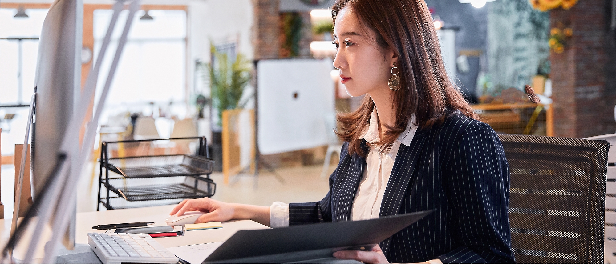 A woman in a blazer sits at a desk, working on a computer with papers in hand, in a bright, modern office space.