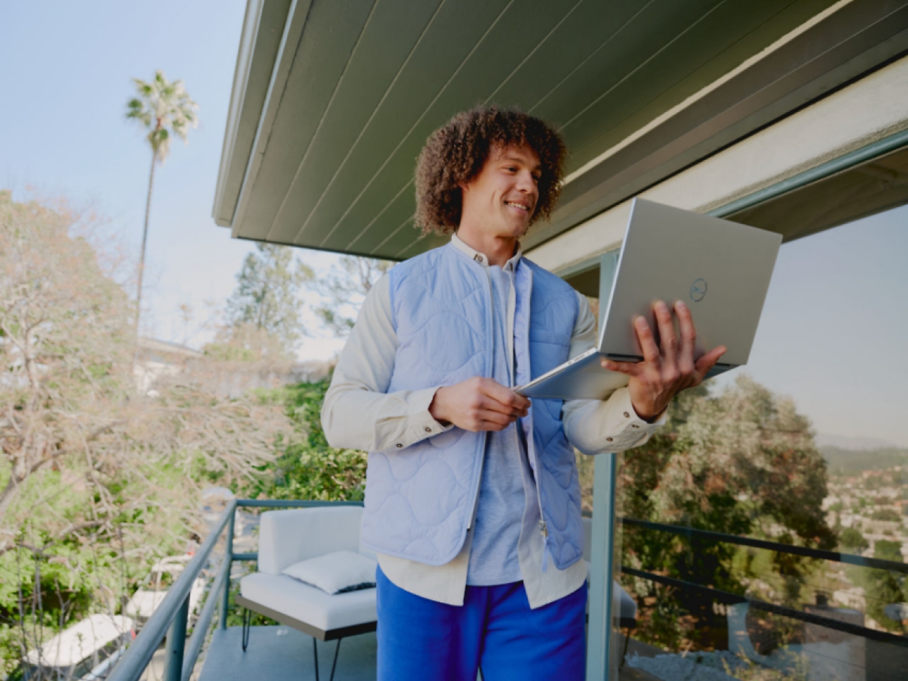 A person stands on a balcony holding an open laptop, smiling.