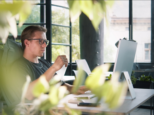 A man with glasses sits at a desk, holding a piece of paper, surrounded by greenery, 